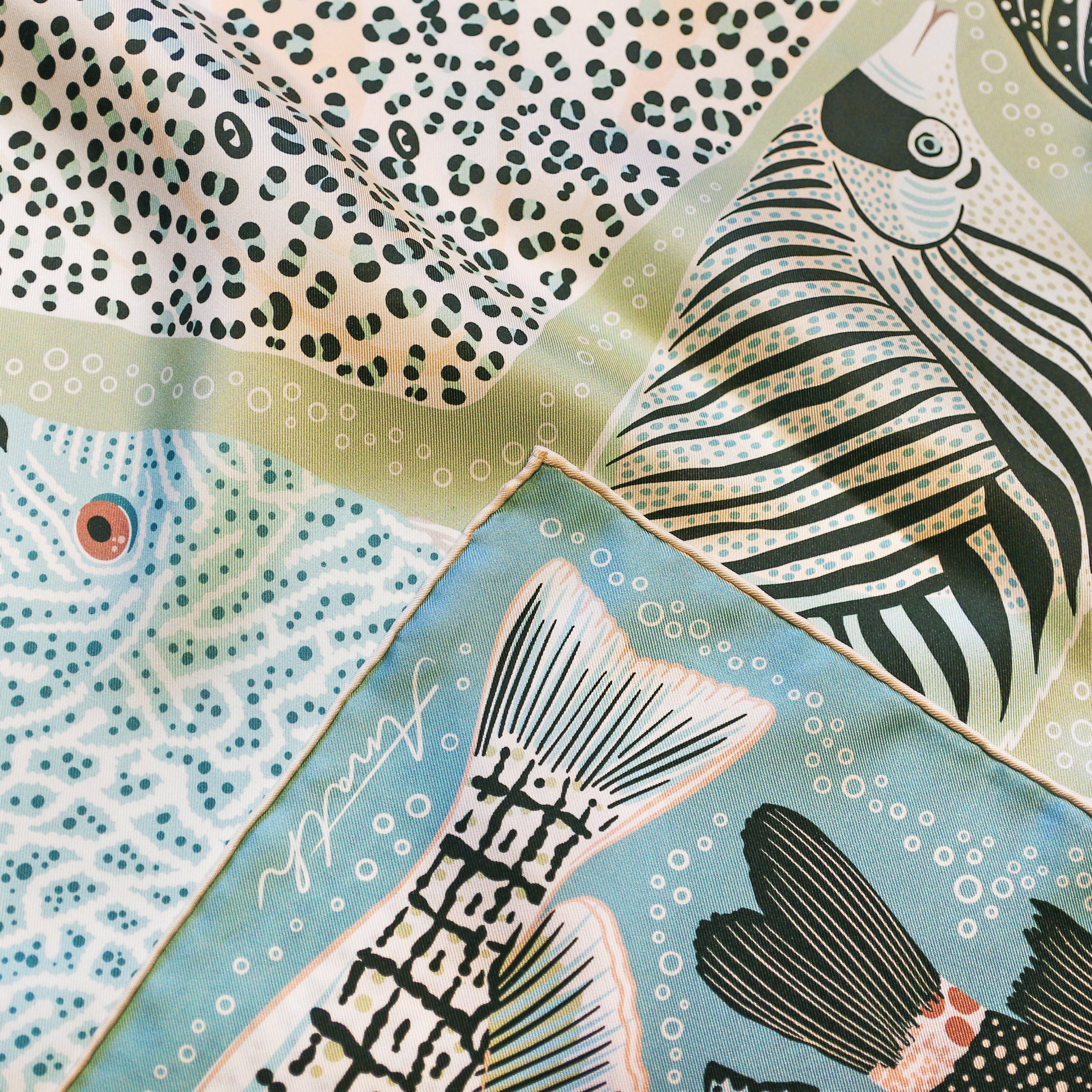 Detail of the Pesce Acqua double-sided silk scarf showing hand-painted fish illustrations and the hand-rolled hem on 100% silk twill.