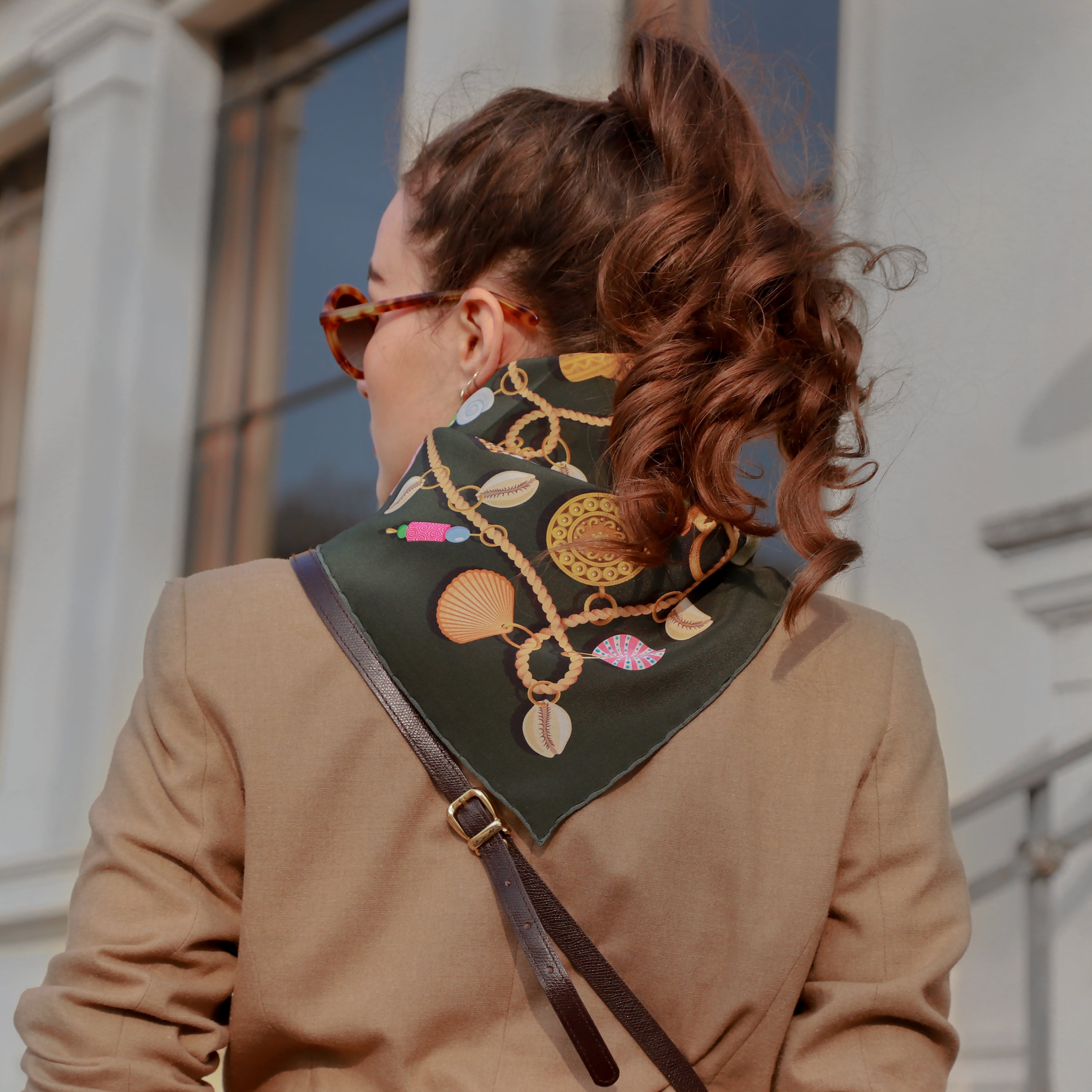 Model wearing the Kauri Delphine silk bandana scarf styled around the neck with a camel coat.