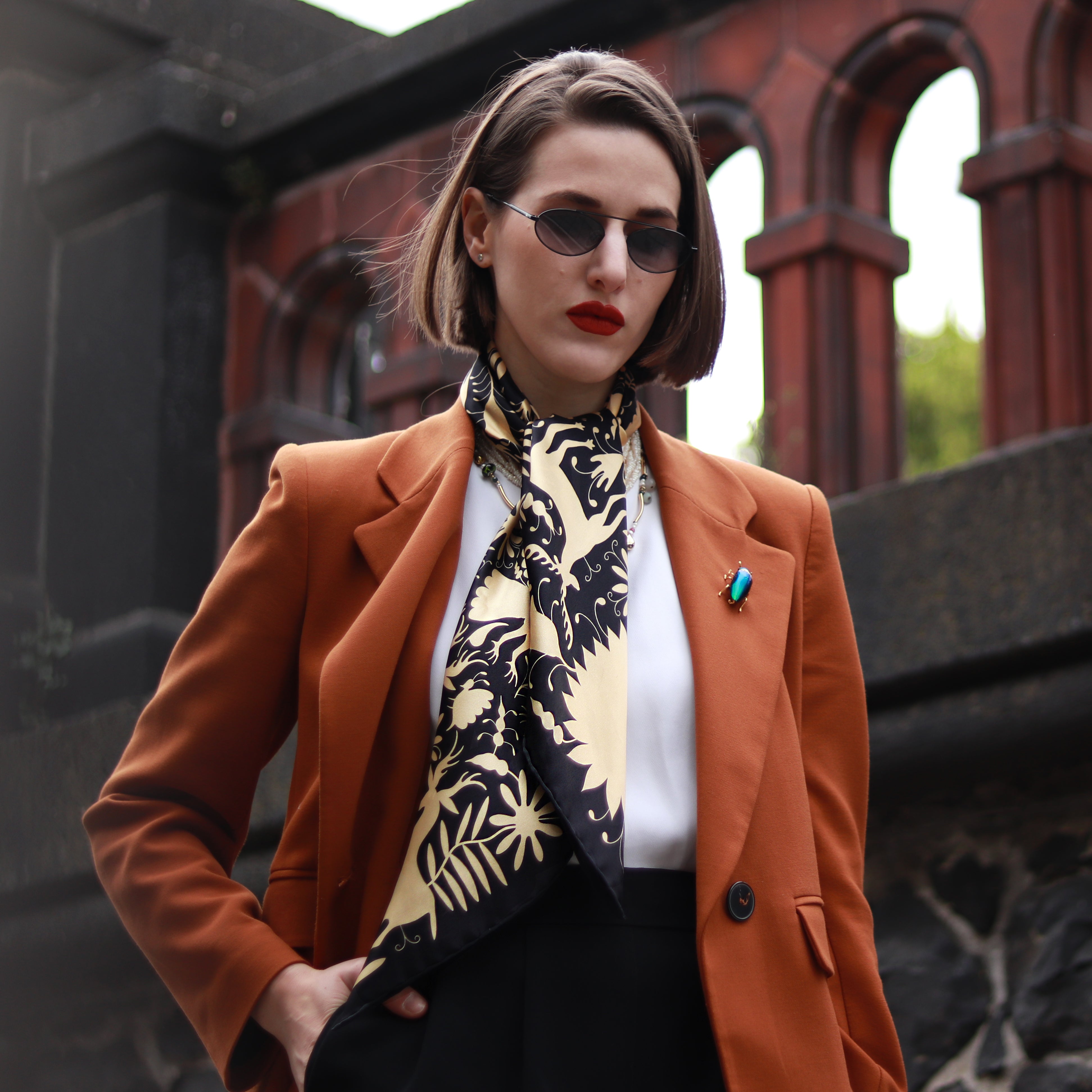 Model wearing the Noé Gold silk scarf styled around the neck with a rust-coloured blazer.