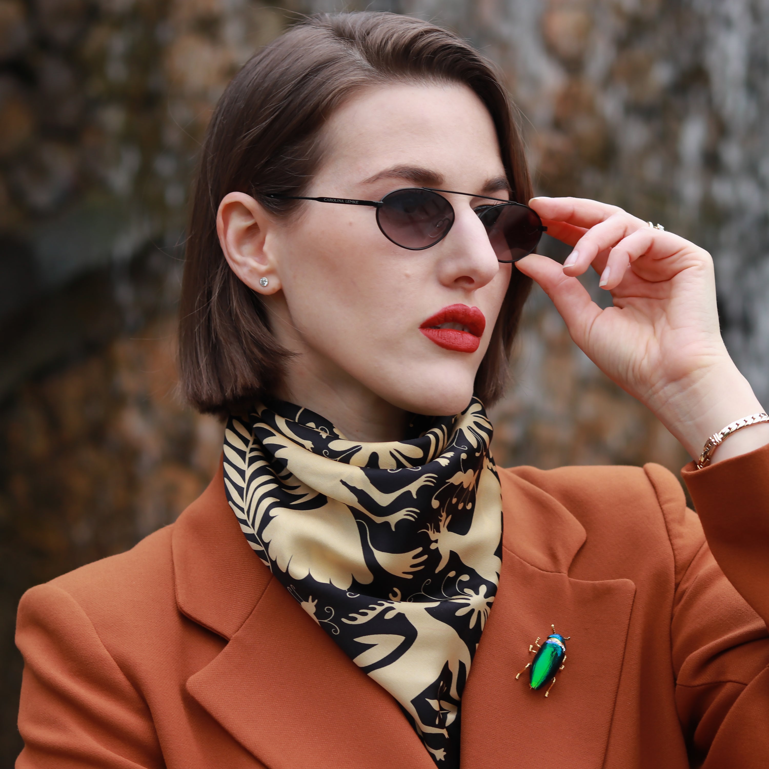 Model wearing the Noé Gold silk scarf styled around the neck with a rust-coloured blazer and sunglasses.