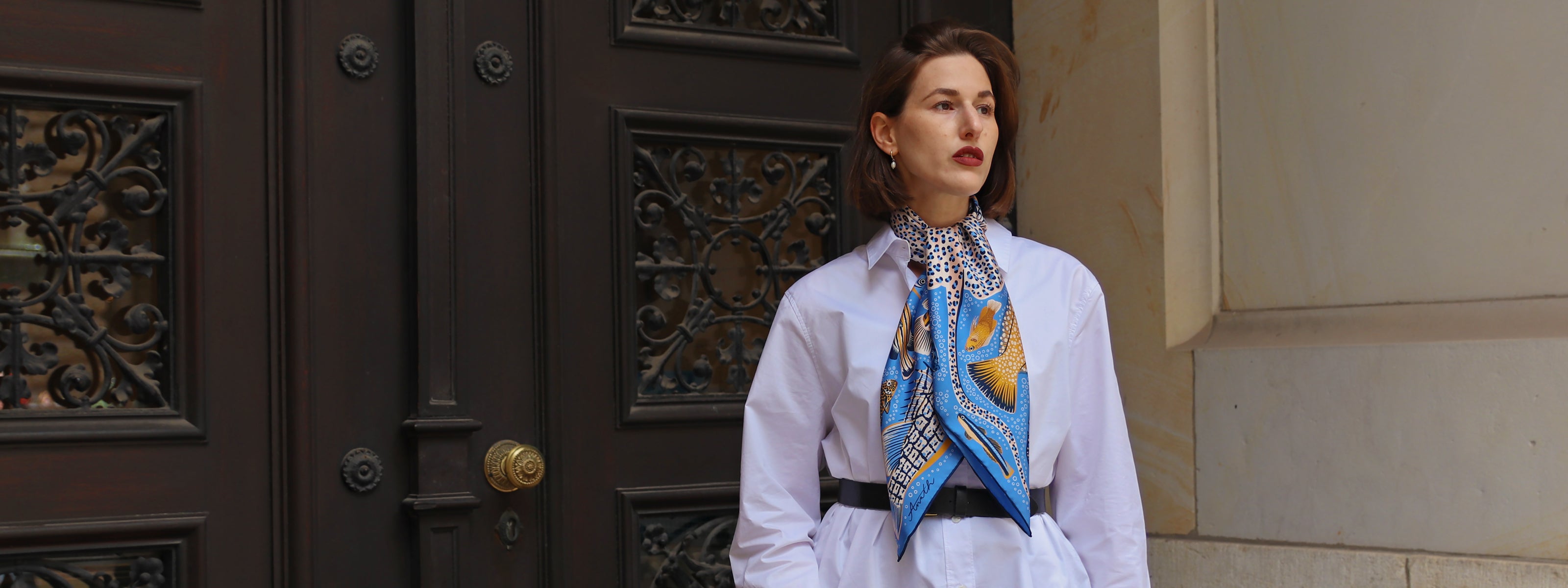 Model wearing the Pesce Iris double-sided silk scarf styled around the neck with cobalt blue and warm gold fish motifs.