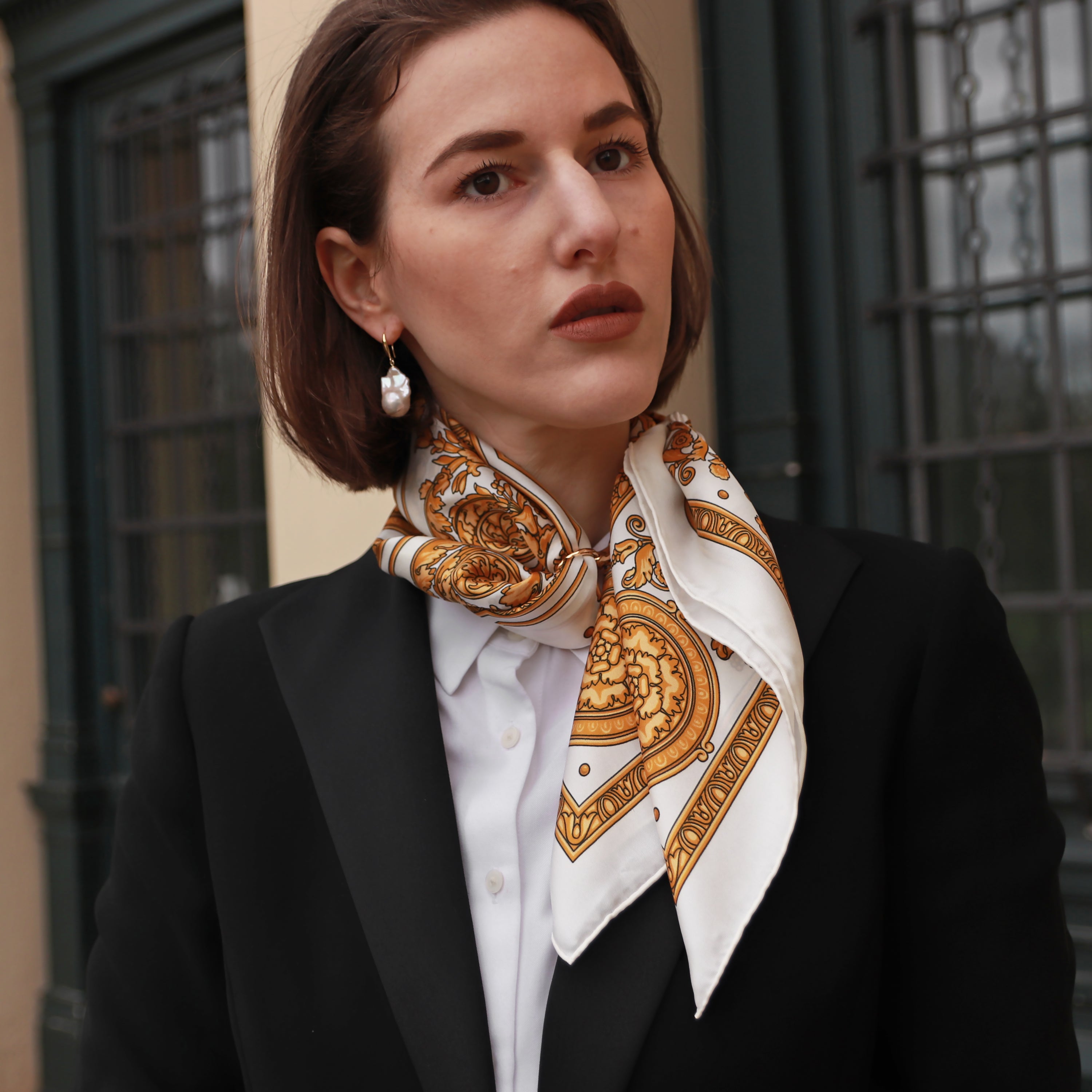 Model wearing the Rococo Natural silk scarf styled around the neck with a black blazer and white shirt.