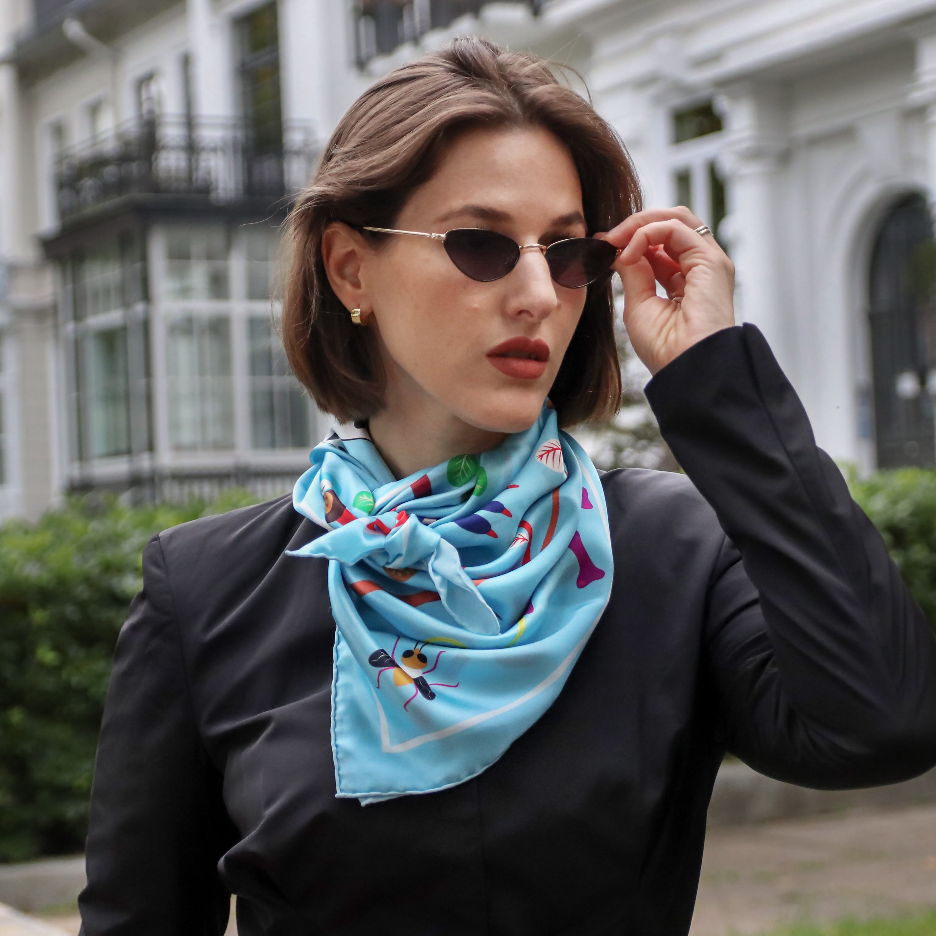Model wearing the Abomey Maya silk scarf styled around the neck with a black blouse and sunglasses.