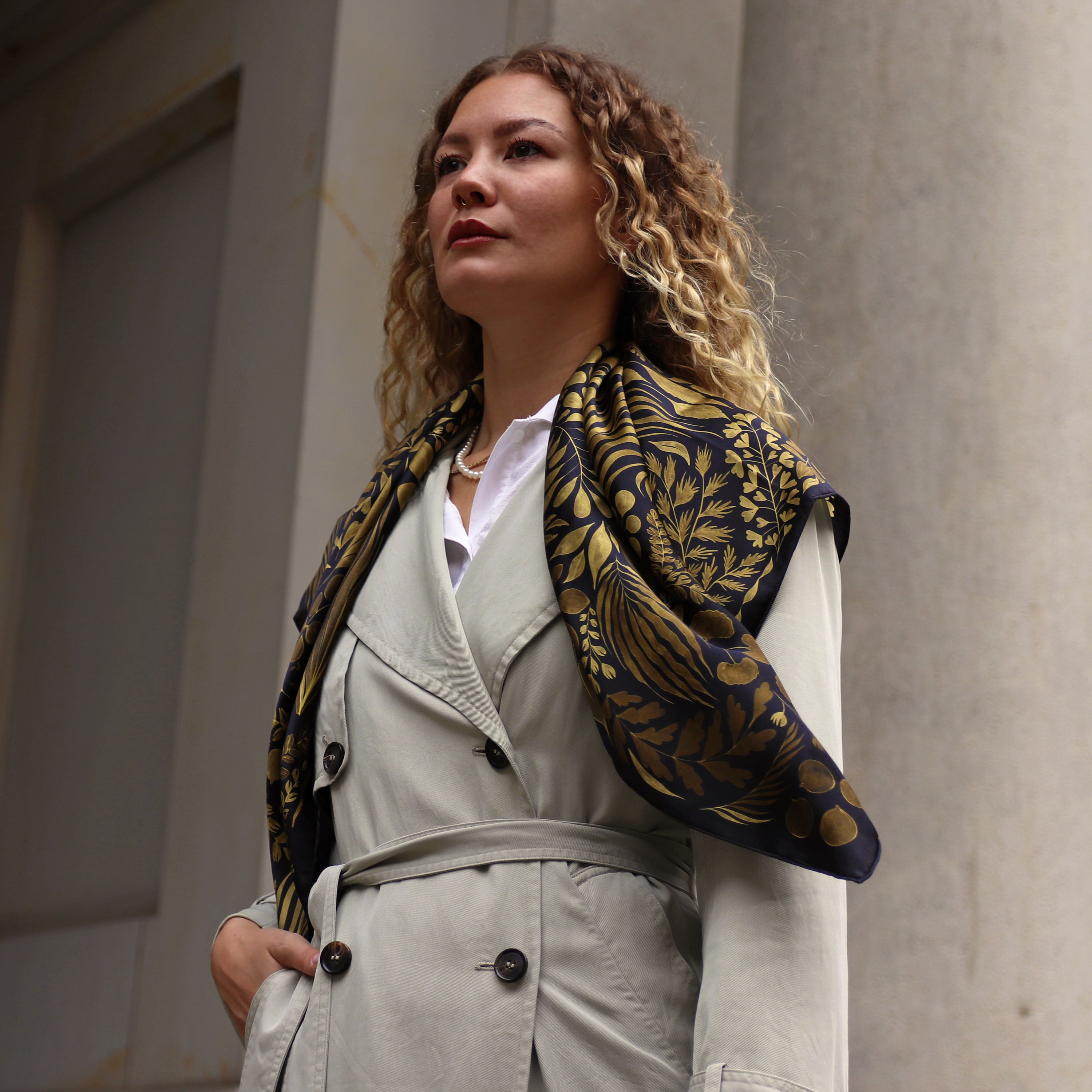 Model wearing the Foglie d'Oro silk scarf draped over the shoulders with a trench coat.