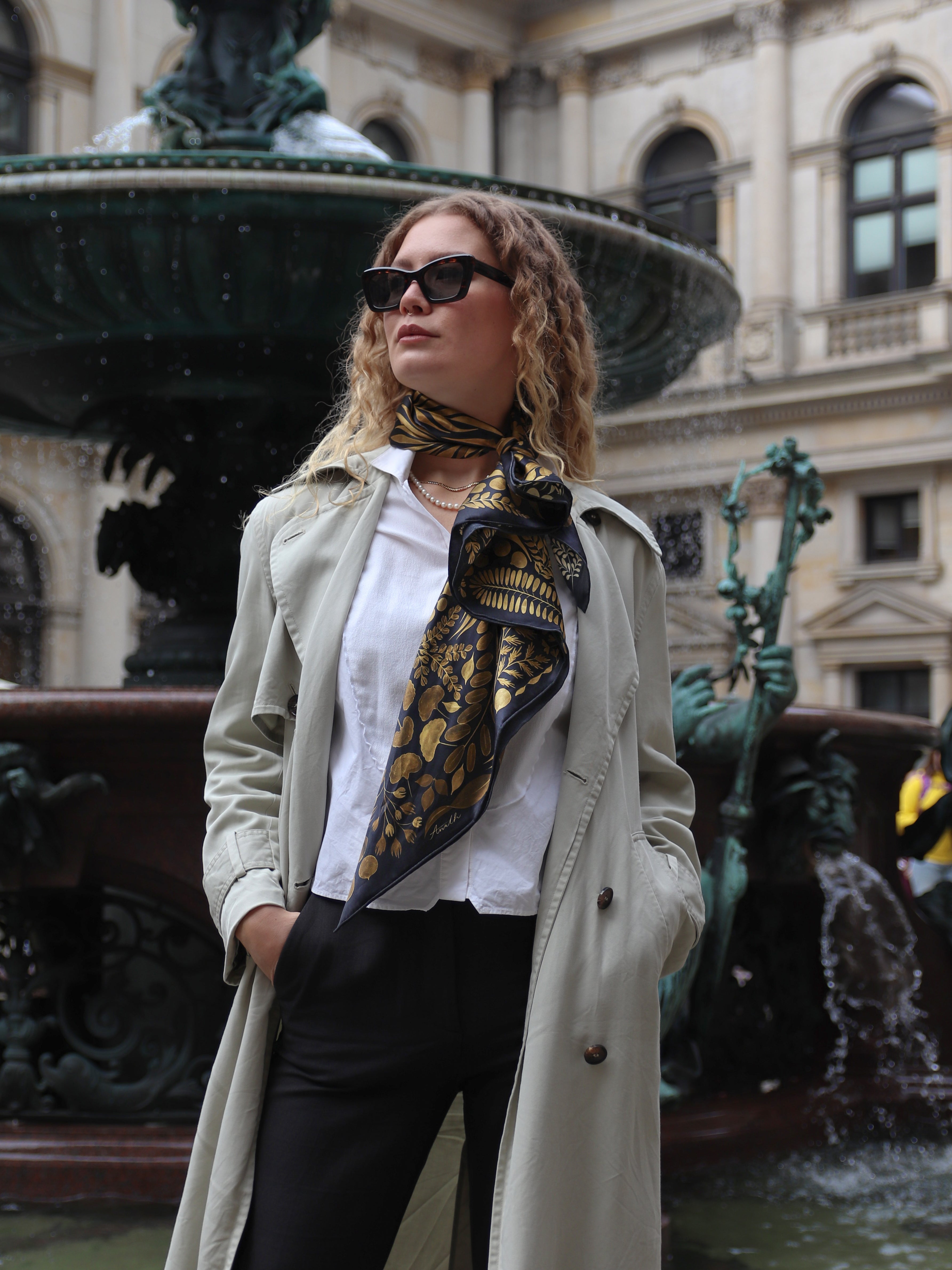 Model wearing the Foglie d'Oro silk scarf styled around the neck with a light trench coat.