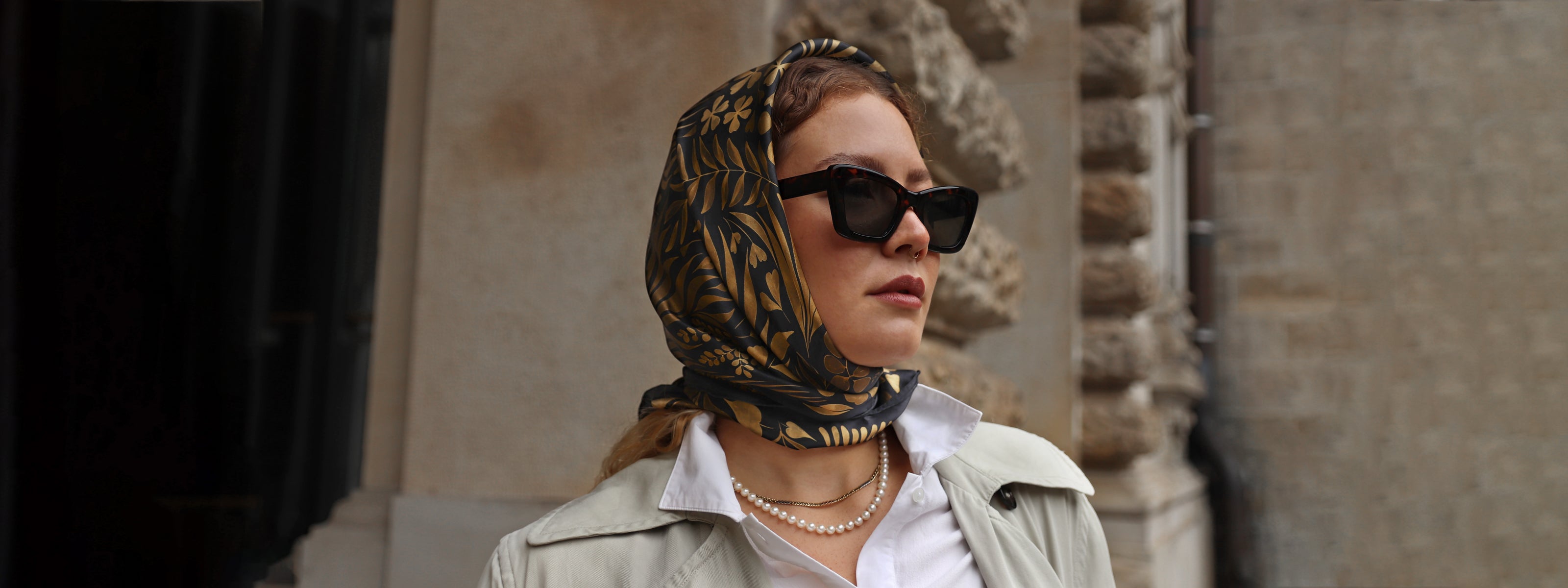 Model wearing the Foglie d'Oro silk scarf styled as a headscarf with a white shirt, pearl necklace and sunglasses.